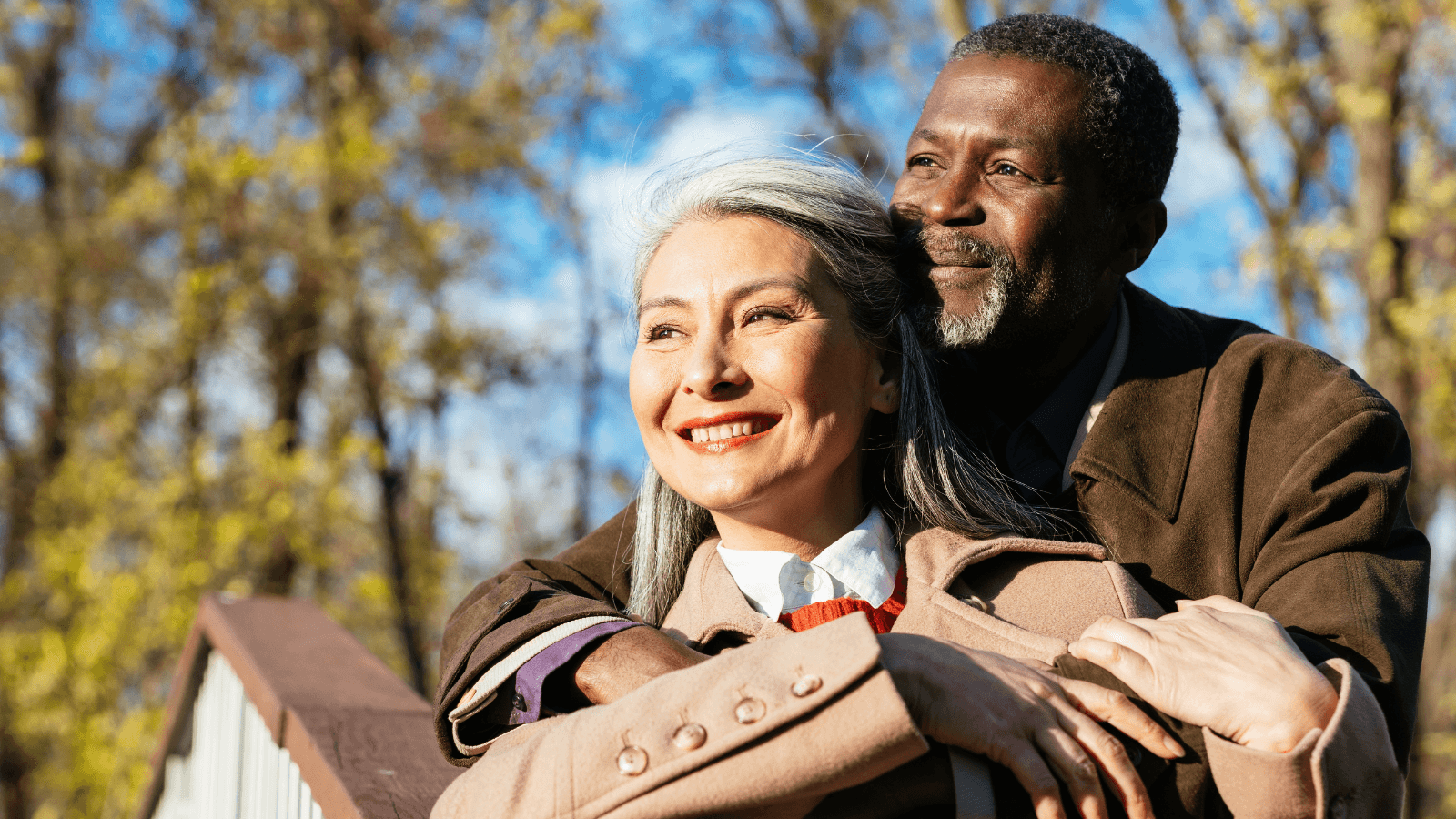 An older couple stands outdoors on a sunny day. The man is embracing the woman from behind as they both smile, with trees and blue sky in the background.