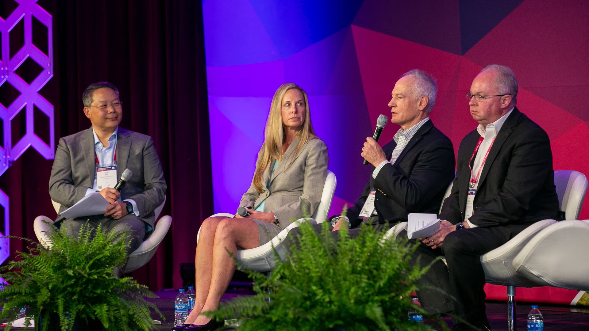 Four people in business attire are seated on a stage panel at the PERA Conference; one man speaks into a microphone while the others listen.
