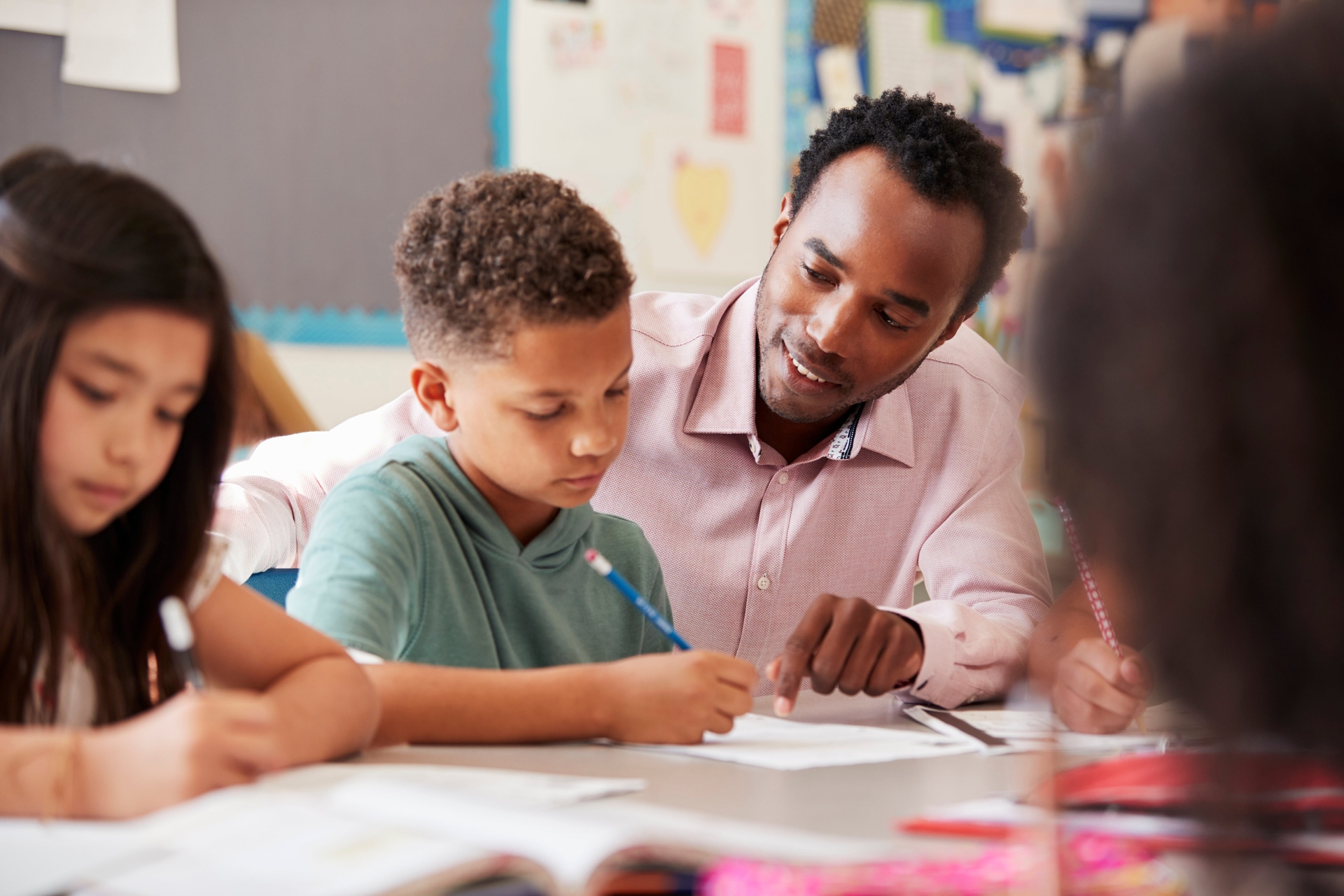 A teacher sits at a table with a group of young students, helping a boy with his schoolwork as others write in their notebooks—dedicated to shaping futures, just like the security provided by a PERA pension.