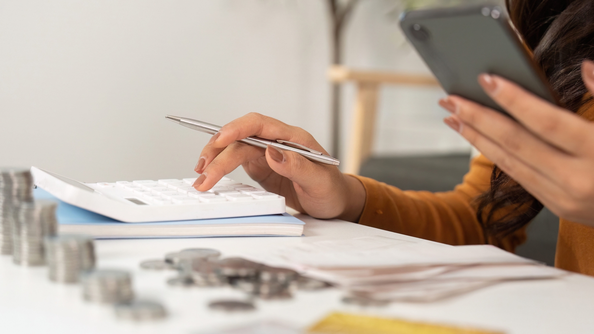 A person uses a calculator and holds a smartphone at a desk with coins, papers, and a few stacked notebooks.