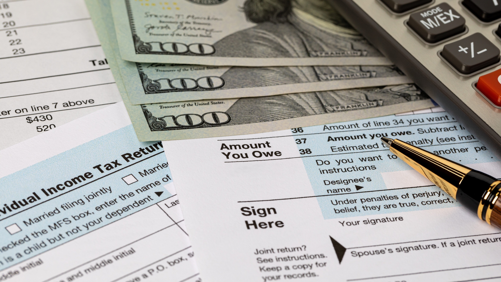 A calculator, pen, and several hundred-dollar bills rest on U.S. individual income tax return forms.