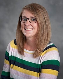 Woman with straight, light brown hair, glasses, and striped sweater in yellow, green, black, and white poses smiling against a gray background.