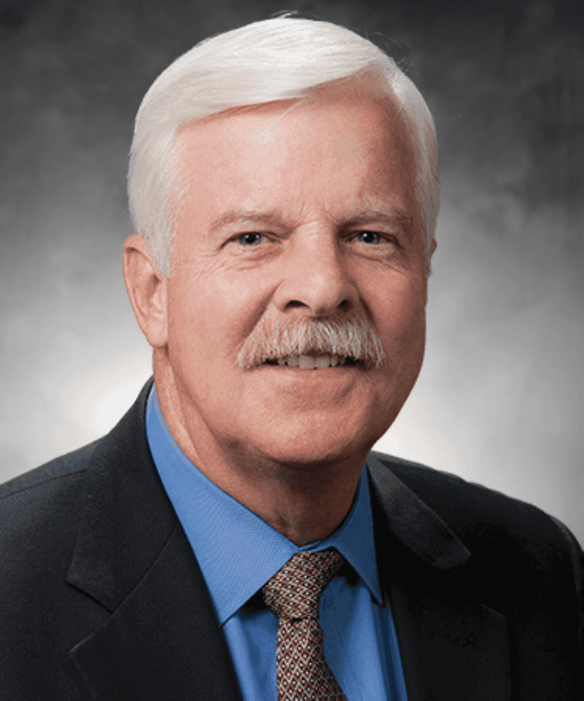 A man with short white hair and a mustache, wearing a dark suit, blue shirt, and patterned tie, poses in front of a neutral grey background.
