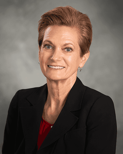 A person with short, light brown hair, wearing a black blazer and red top, smiling in front of a gray background.