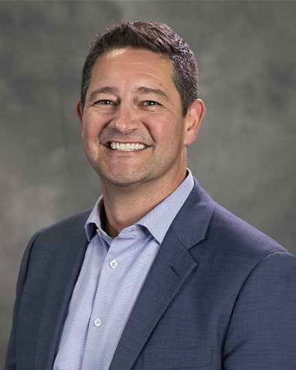 A man in a blue suit jacket and light blue shirt smiles at the camera in front of a gray background.