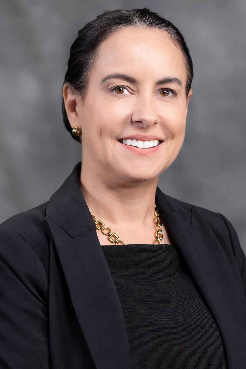 Woman in a black blazer and gold necklace smiles at the camera against a gray background.