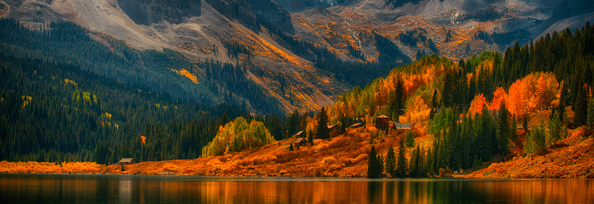Colorful autumn trees and rustic wooden cabins line the shore of a calm lake, set against a backdrop of forested mountains.