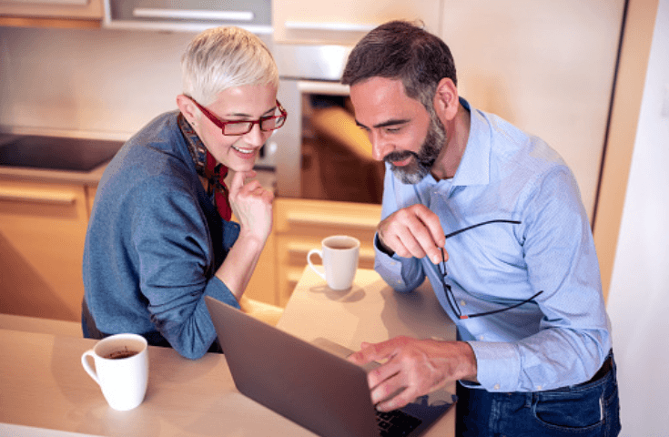 Two adults sit at a kitchen counter with coffee mugs, looking at a laptop screen and smiling while having a discussion.