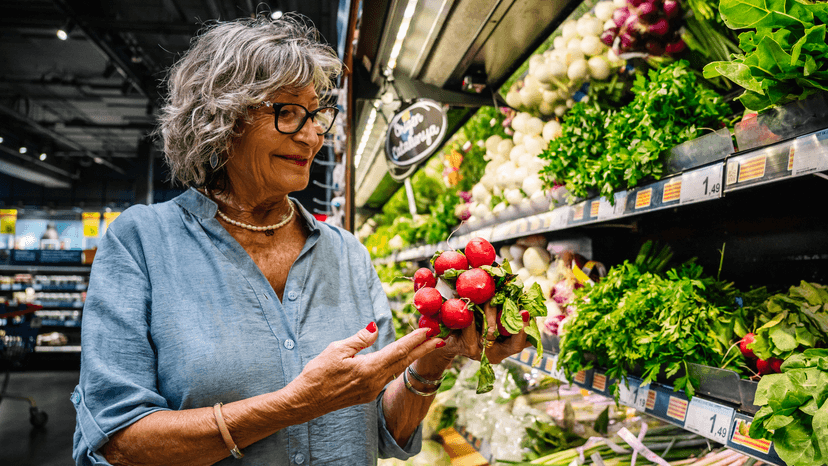 A gray-haired woman holding a bunch of radishes in a supermarket produce aisle.