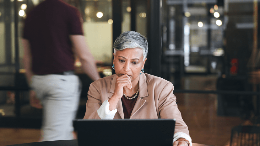A woman with short gray hair sits at a table, looking at a laptop with a thoughtful expression. A person walks by in the background inside a modern office space.