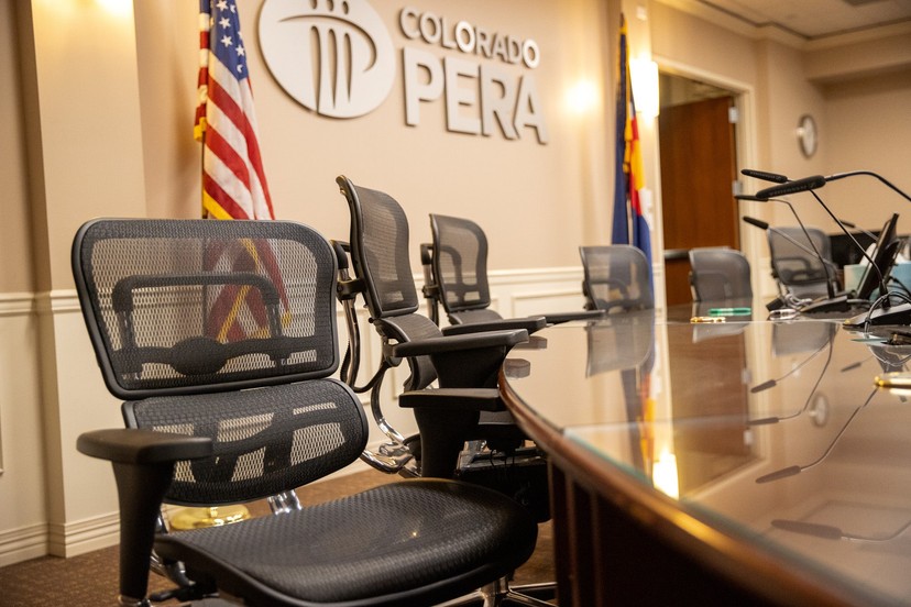 An empty conference room with mesh office chairs around a polished wooden table. The wall features the logo for Colorado PERA. An American flag and a Colorado state flag are in the background.