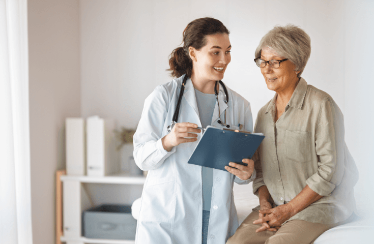 A doctor holding a clipboard talks to an older woman sitting on an exam table in a medical office.
