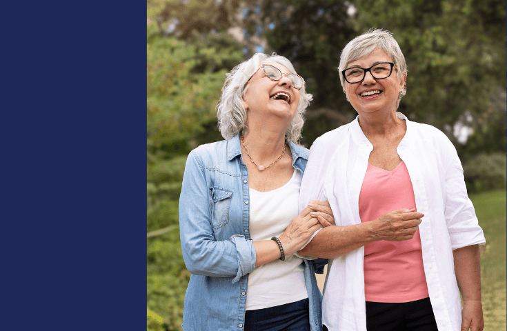 Two older women, one in a blue shirt and the other in a white shirt, walk outdoors arm-in-arm, smiling and laughing with greenery in the background.