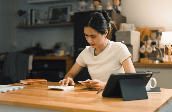 Woman sits at a kitchen table using a calculator and smartphone, with a tablet, a cup, and books nearby.
