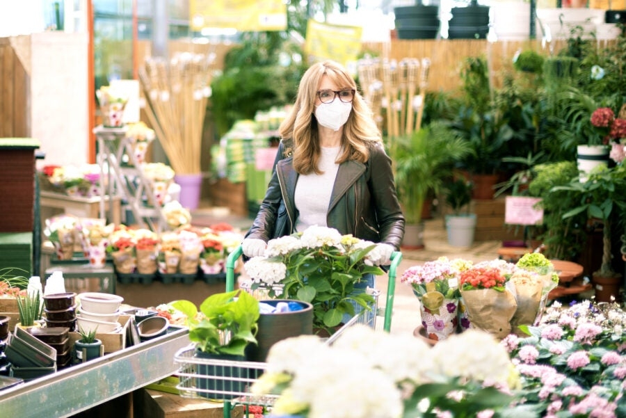 A woman wearing a mask and glasses shops for plants in a garden center, surrounded by various flowers and gardening supplies.