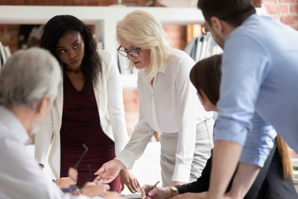 A group of professionals having a discussion around a table in an office setting. A woman with blonde hair is speaking and gesturing, while others listen attentively.