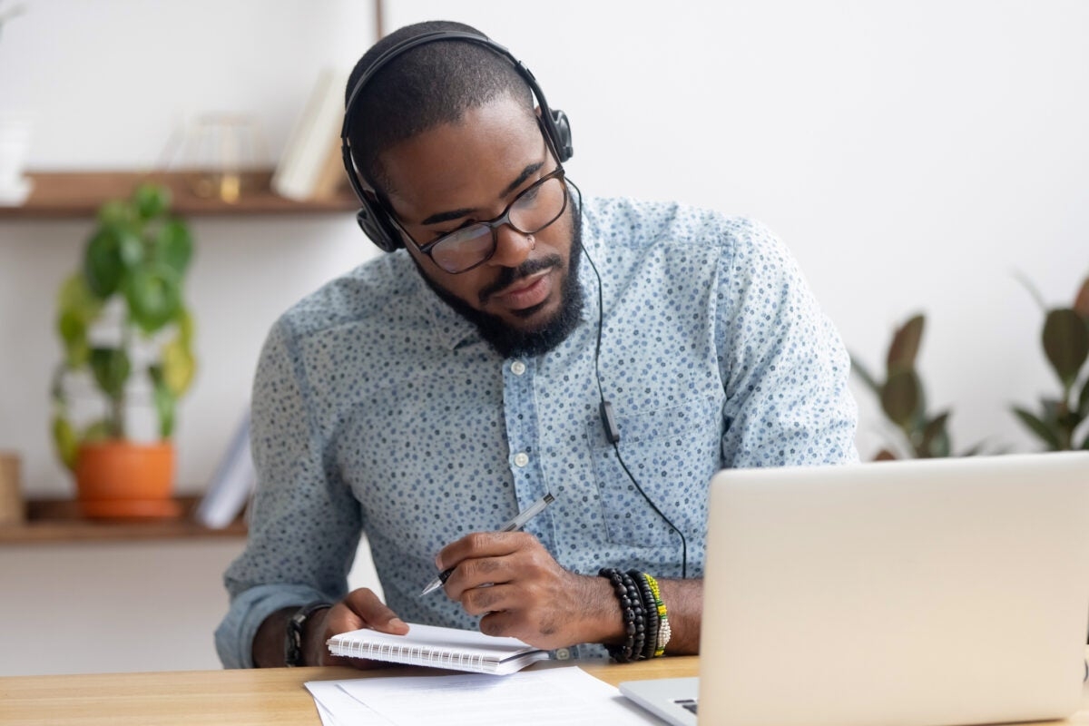 A person wearing headphones and glasses, taking notes in a notebook while using a laptop, seated at a desk with plants in the background.