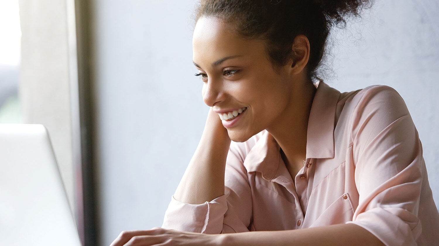 A woman with a smile is seated at a table focusing on a laptop in front of her, wearing a light pink blouse.