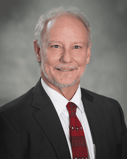 A man with gray hair and a beard wearing a dark suit, white shirt, and red patterned tie is smiling at the camera against a neutral background.