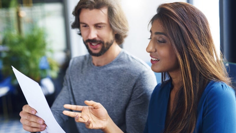 Two people in a well-lit room are engaged in discussion while looking at a sheet of paper. The foreground shows a man with a beard and a woman with long hair.