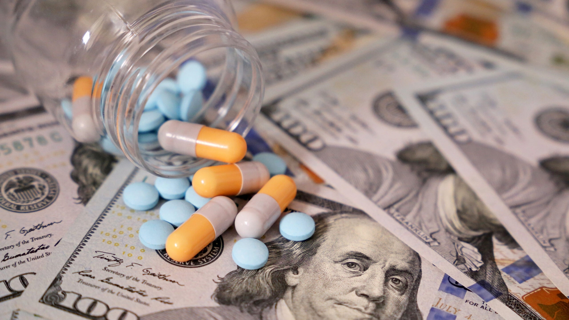 A pill bottle with various types of pills spilling out of it sits atop a pile of $100 bills.