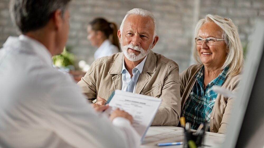An older couple sits across from a doctor who is holding documents, discussing something in a medical office setting.