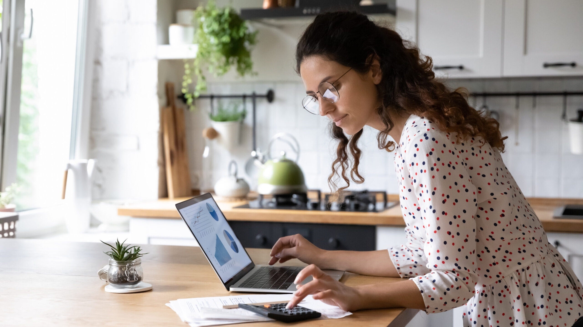 A woman standing at her kitchen counter using a laptop and calculator.