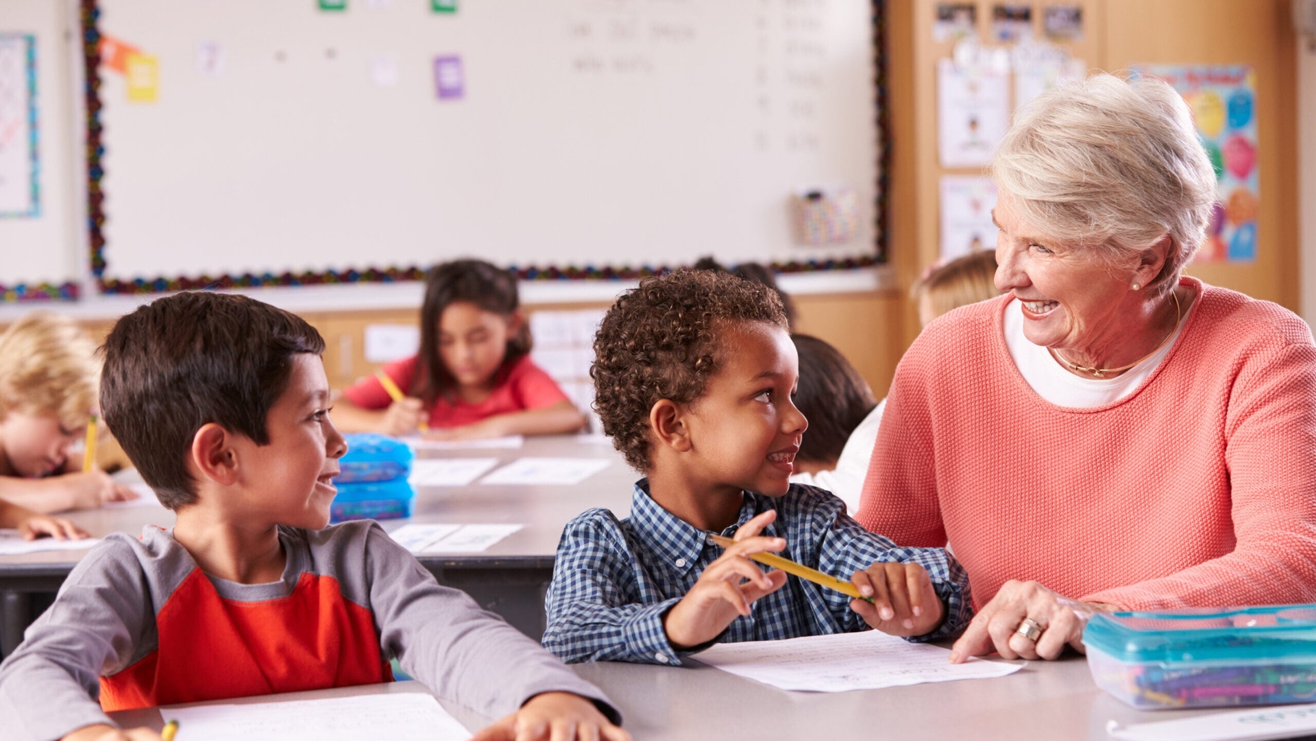 An older woman sits at a desk in a classroom, smiling and talking with two young boys while other children work in the background, showing the positive financial impact of working after retirement.