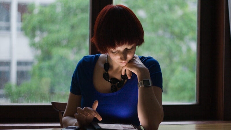 Woman sitting in a dark room with a tablet illuminating her face, a window behind her has blurred trees.