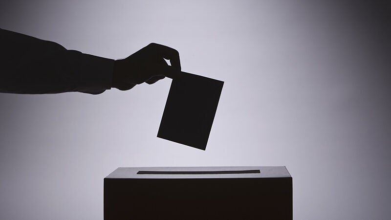 A silhouette of an arm, paper, and box, giving the illusion of someone casting a vote.