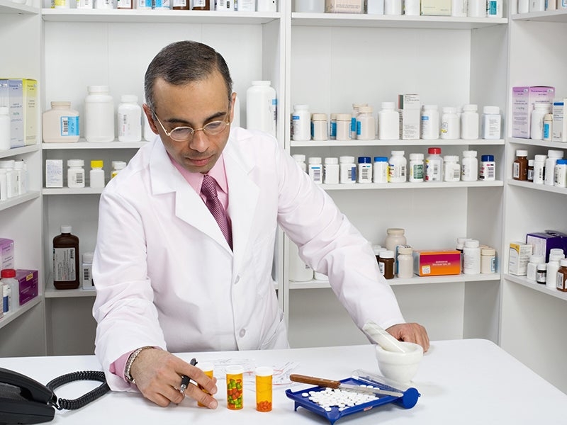 A pharmacist in a white coat prepares and sorts medication in a pharmacy, surrounded by shelves of medicine bottles and a mortar and pestle on the counter.