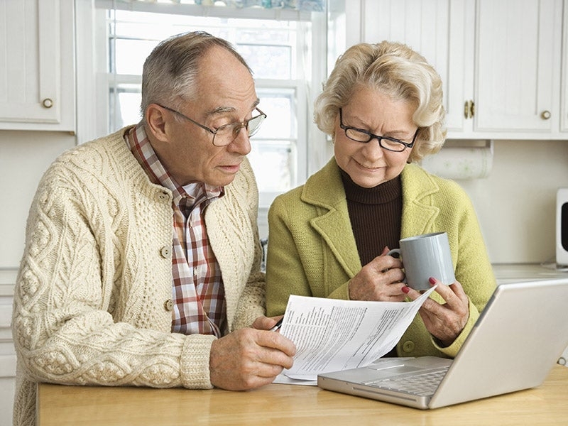 An elderly couple sitting looking at a paper, the woman is holding a coffee mug and a laptop sits open in front of them.
