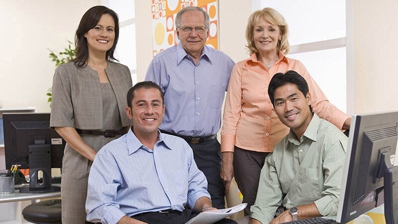 A group of five people in front of computers in a professional setting are smiling at the camera.
