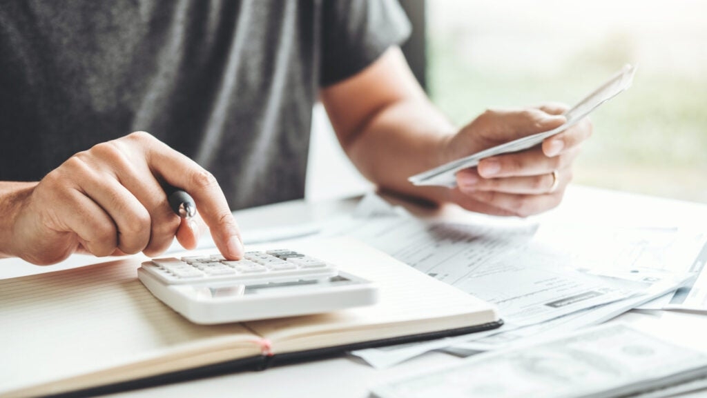 A person uses a calculator while holding a phone, surrounded by papers and a notebook on a desk.