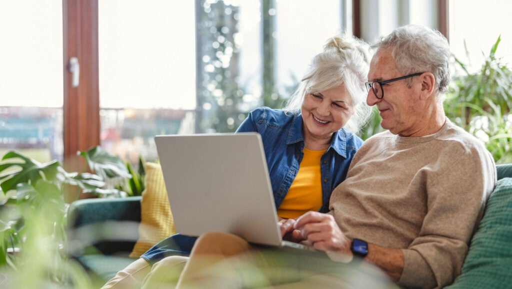 An elderly couple sits on a sofa in a bright room, smiling and looking at a laptop together. They are surrounded by green indoor plants.