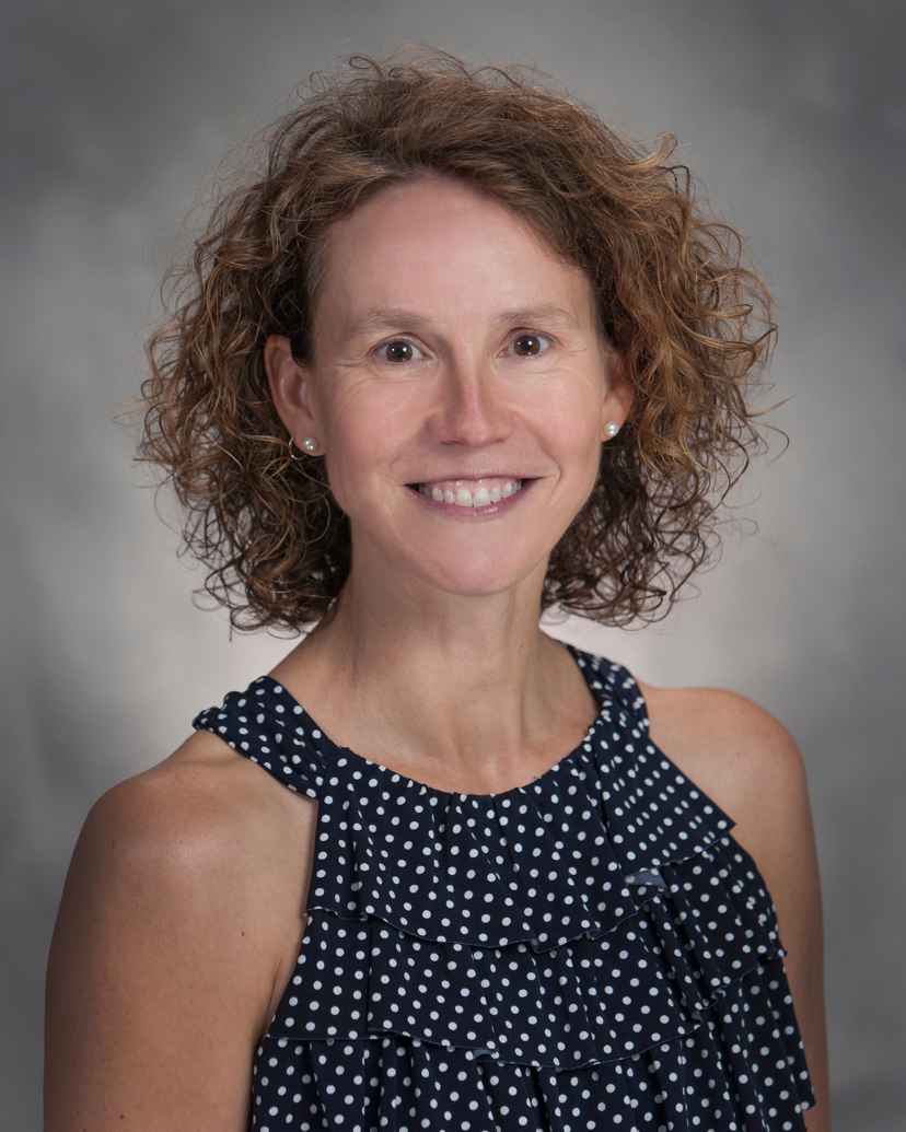 Woman with curly brown hair wearing a sleeveless navy blue and white polka dot blouse, smiling at the camera with a neutral gray background.