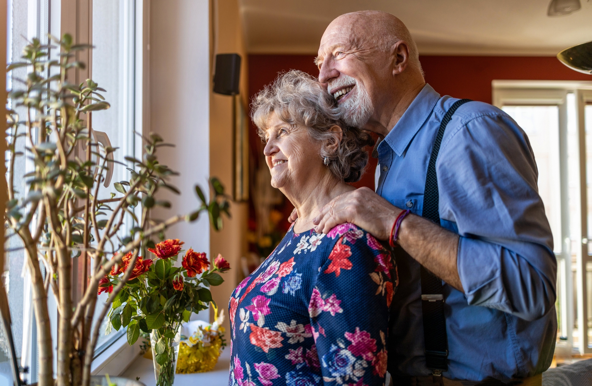 An older couple standing together and looking out a window.