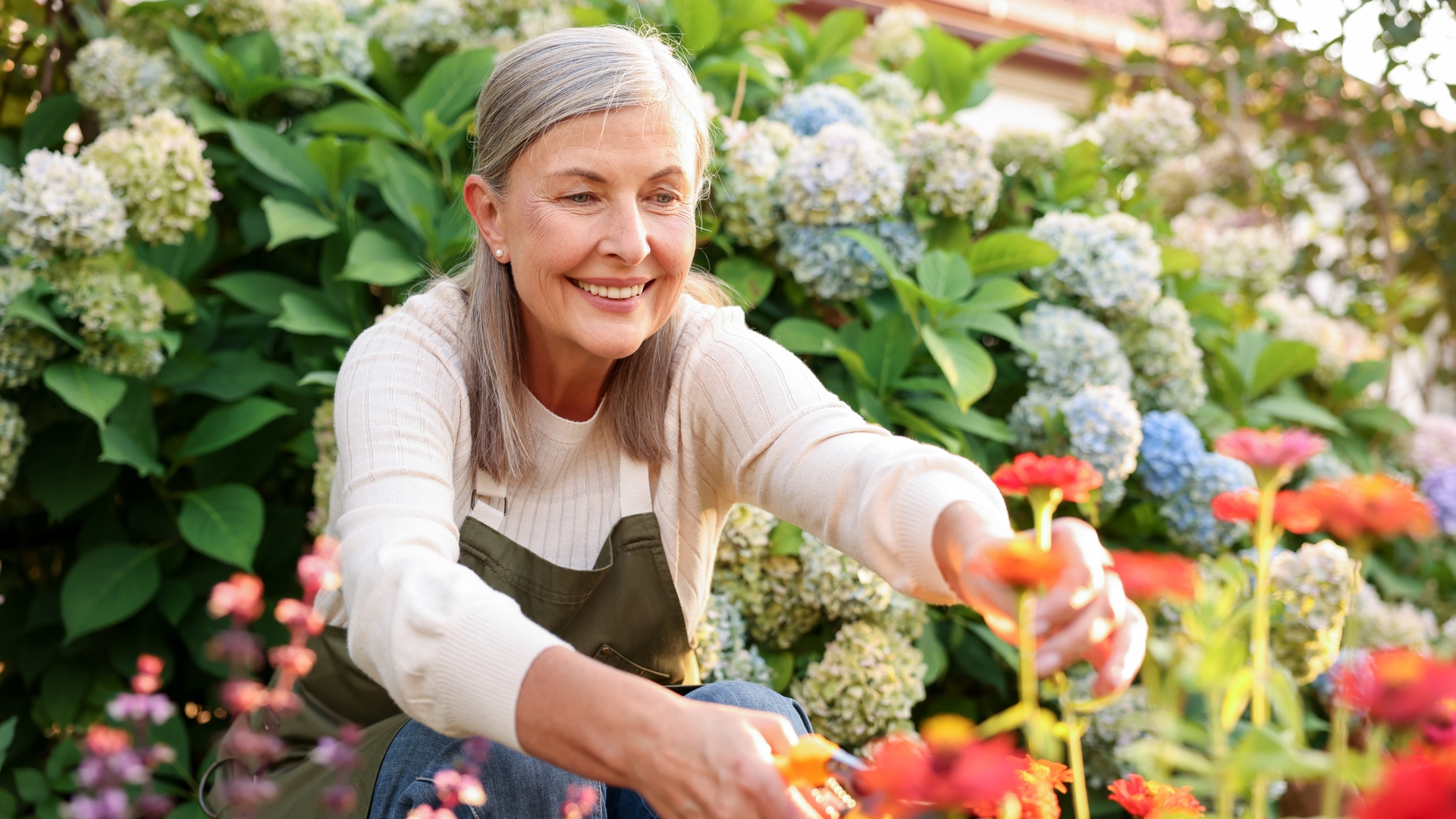 A gray-haired woman kneeling and clipping flowers in her garden.
