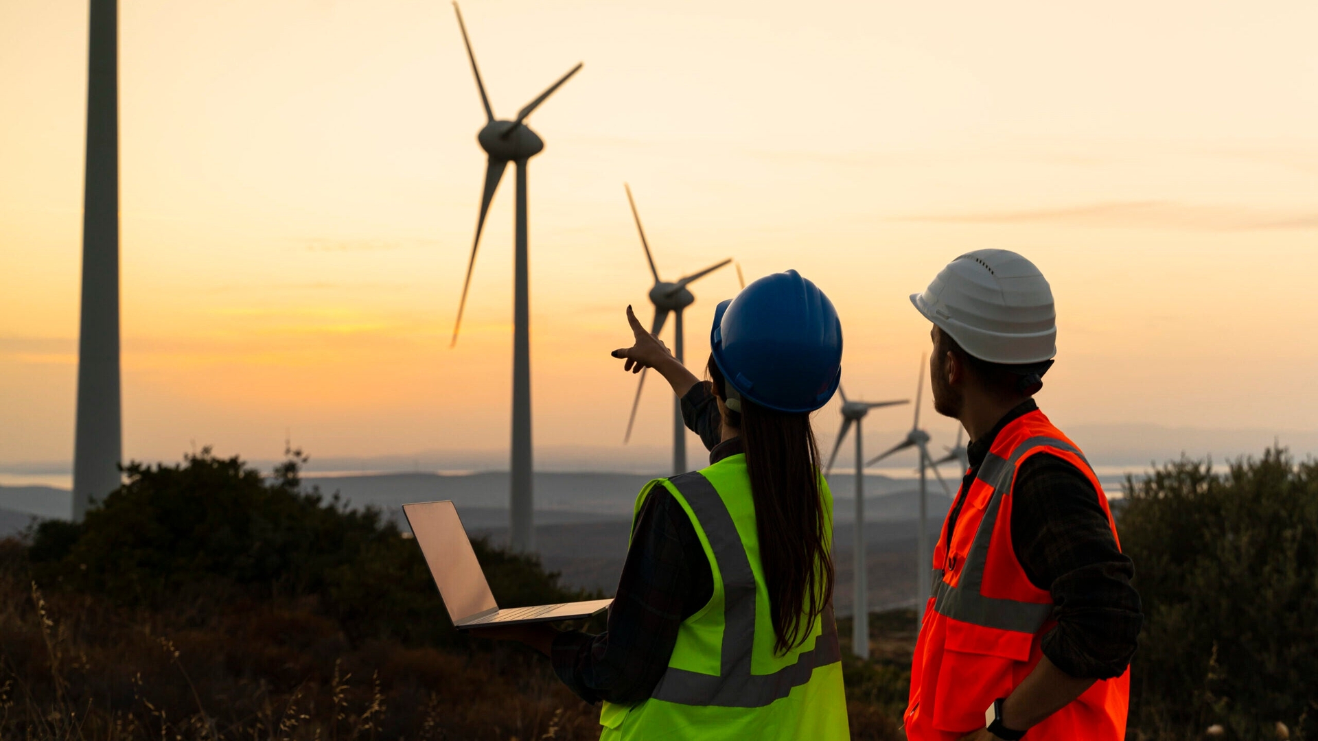 Two workers in safety gear inspect a wind farm with wind turbines at sunset, one holding a laptop and the other pointing towards the turbines.