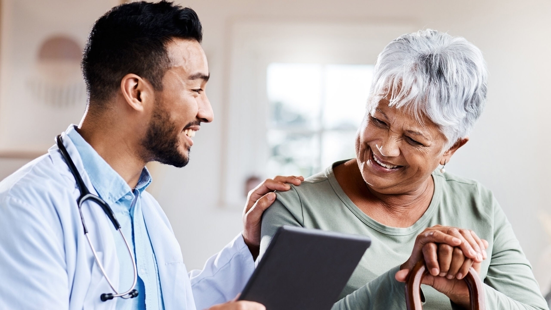 A doctor holding a tablet talks and smiles with an older woman who is holding a cane, sitting across from him in a well-lit room.