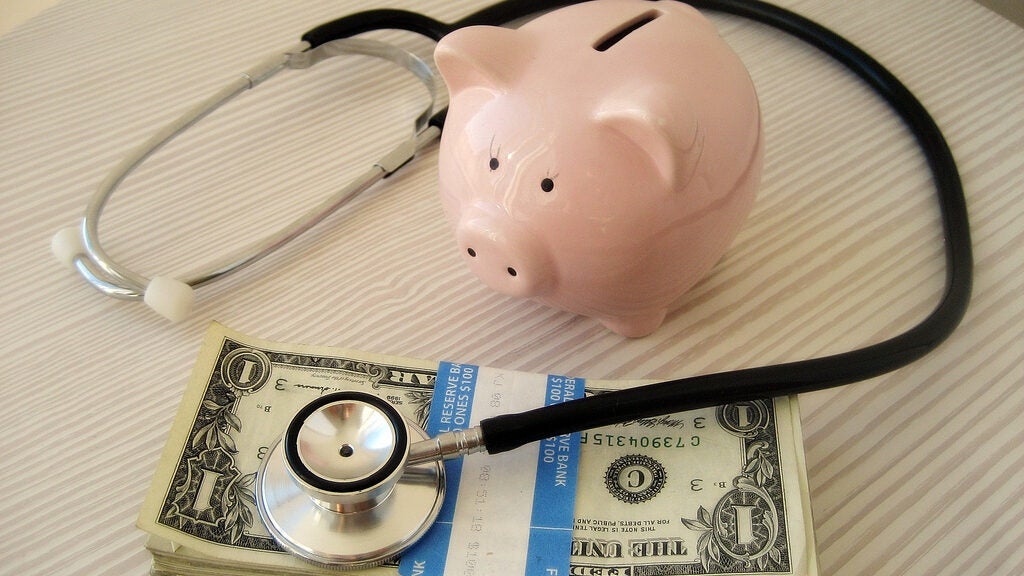 A pink piggy bank, a stethoscope, and a stack of dollar bills on a striped surface.
