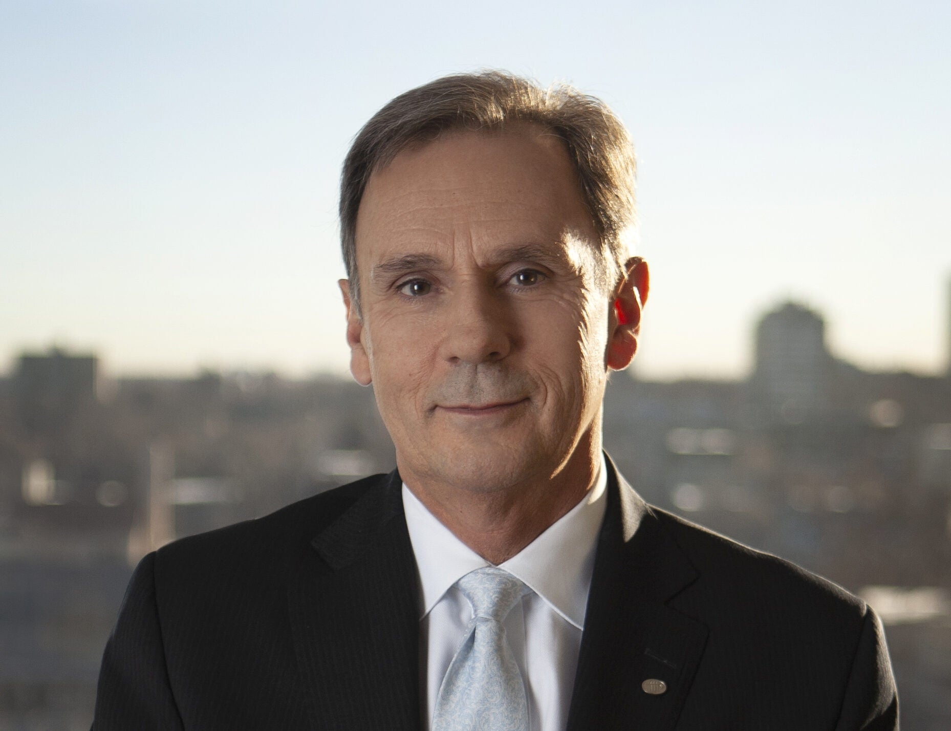 A middle-aged man in a suit and tie poses for a professional portrait, the out-of-focus cityscape behind him subtly hinting at his recent success as PERA board member Ron Baker was reelected.
