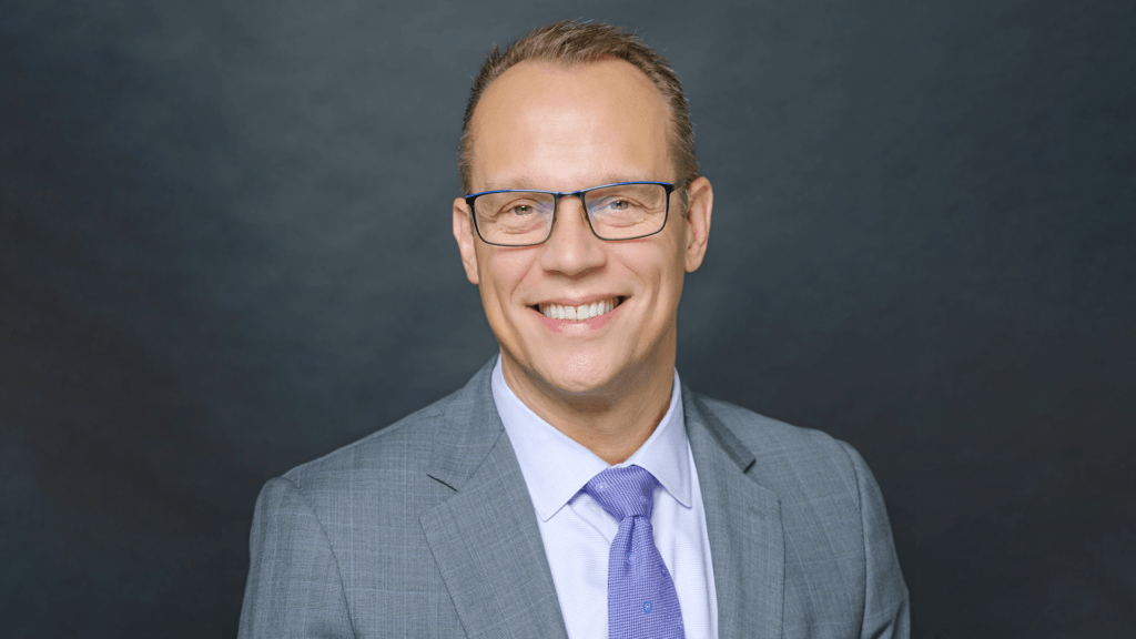 A smiling individual wearing glasses, a gray suit, a light blue shirt, and a blue tie against a dark background.