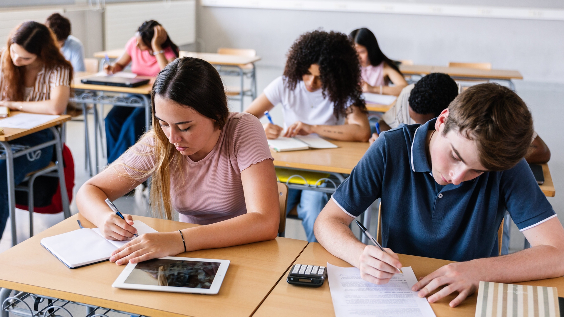 Students sitting at desks in a classroom, writing on paper. Some have tablets and calculators on their desks.